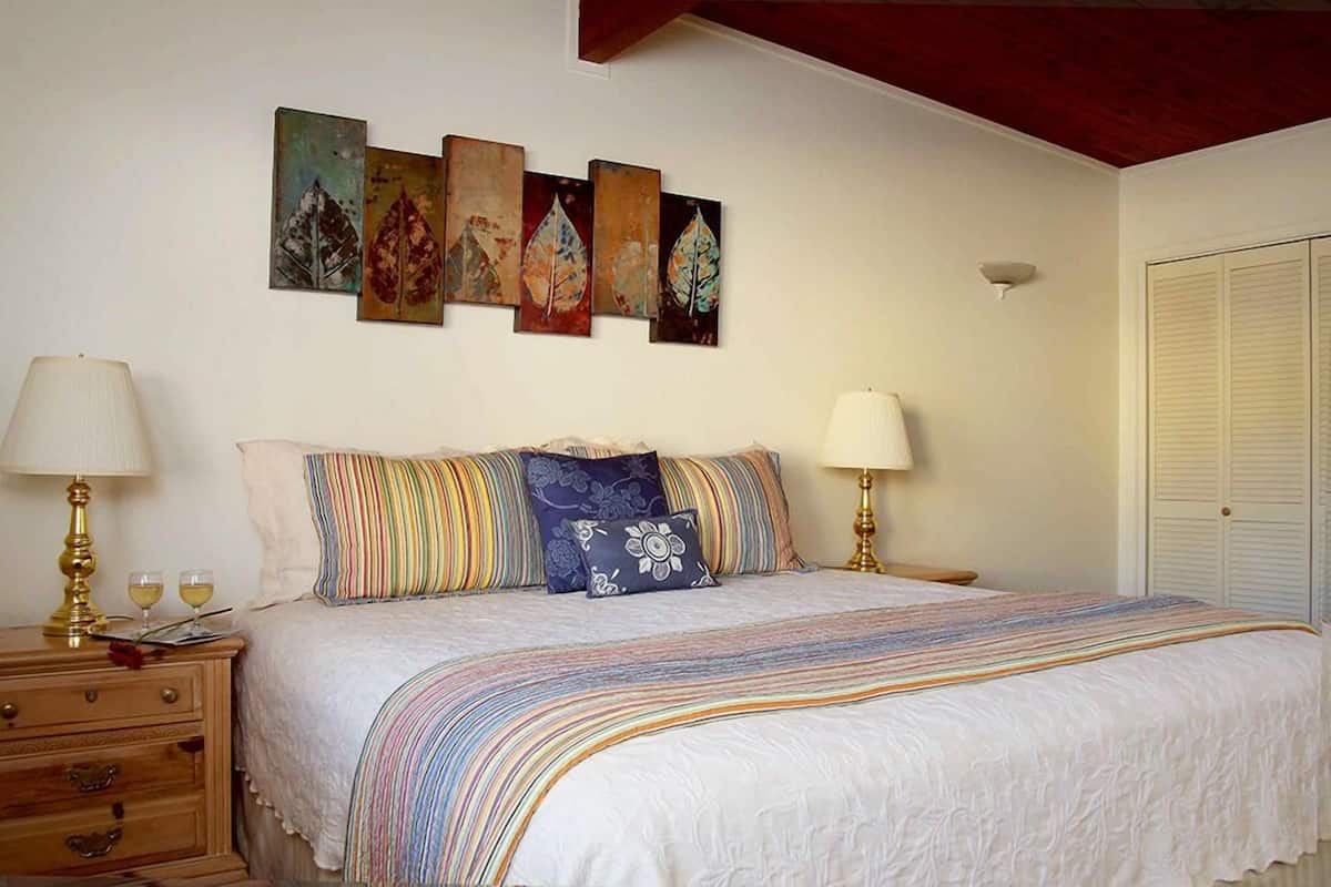 A bedroom with a bed, two bedside tables, a nightstand, a wall-mounted lamp, and a set of four framed leaf paintings.