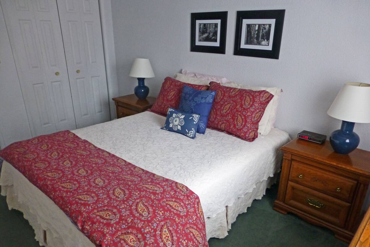 A neatly made bed with a red and white patterned bedspread, two blue lamps on wooden nightstands, and two framed pictures on the wall.