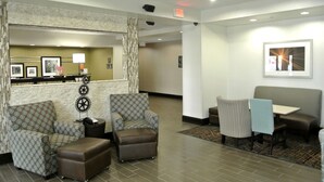 A hotel lobby with a reception desk, patterned armchairs, a small table with a chair, and a wall-mounted picture. - Hampton Inn Carrizo Springs (Carrizo Springs)