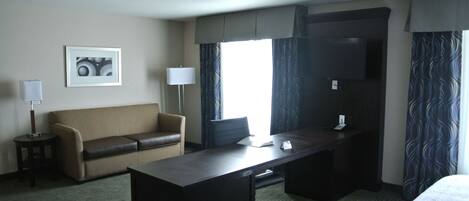 A hotel room with a bed, a desk, a chair, a sofa, a TV, and a window with curtains.
