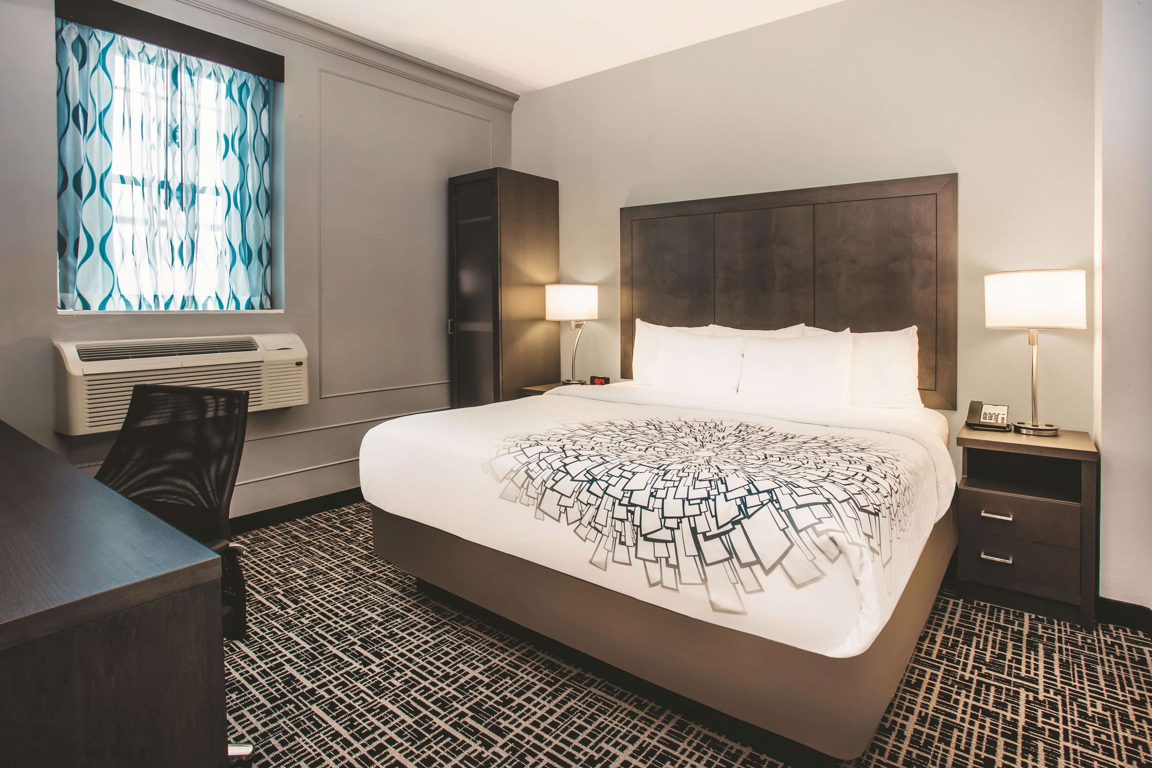 A hotel room with a large bed, a desk, a chair, a window with curtains, a wall-mounted air conditioner, and two bedside lamps.