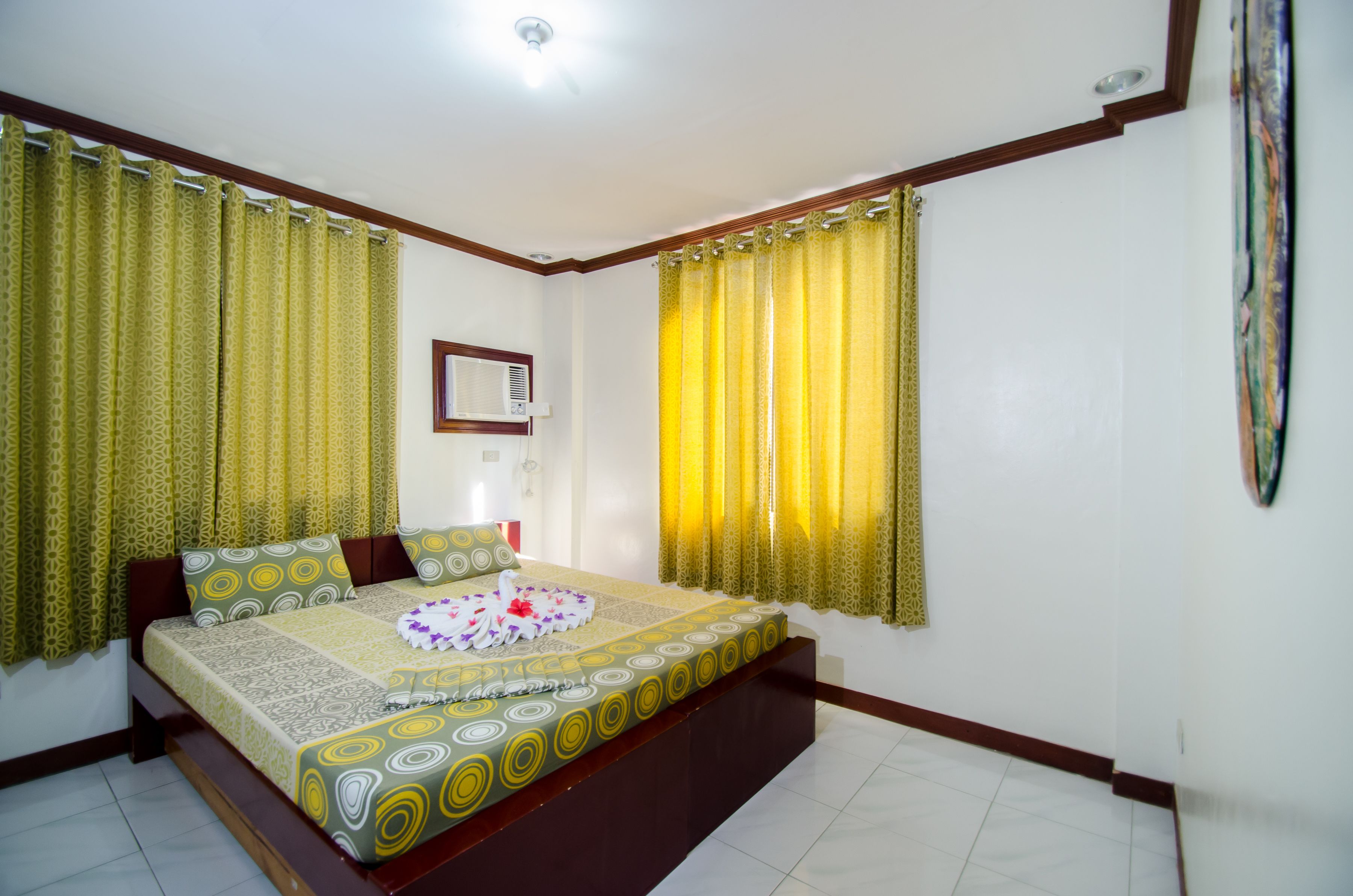 A hotel room with a large bed, yellow patterned curtains, a small bedside table, and a wall-mounted air conditioner.