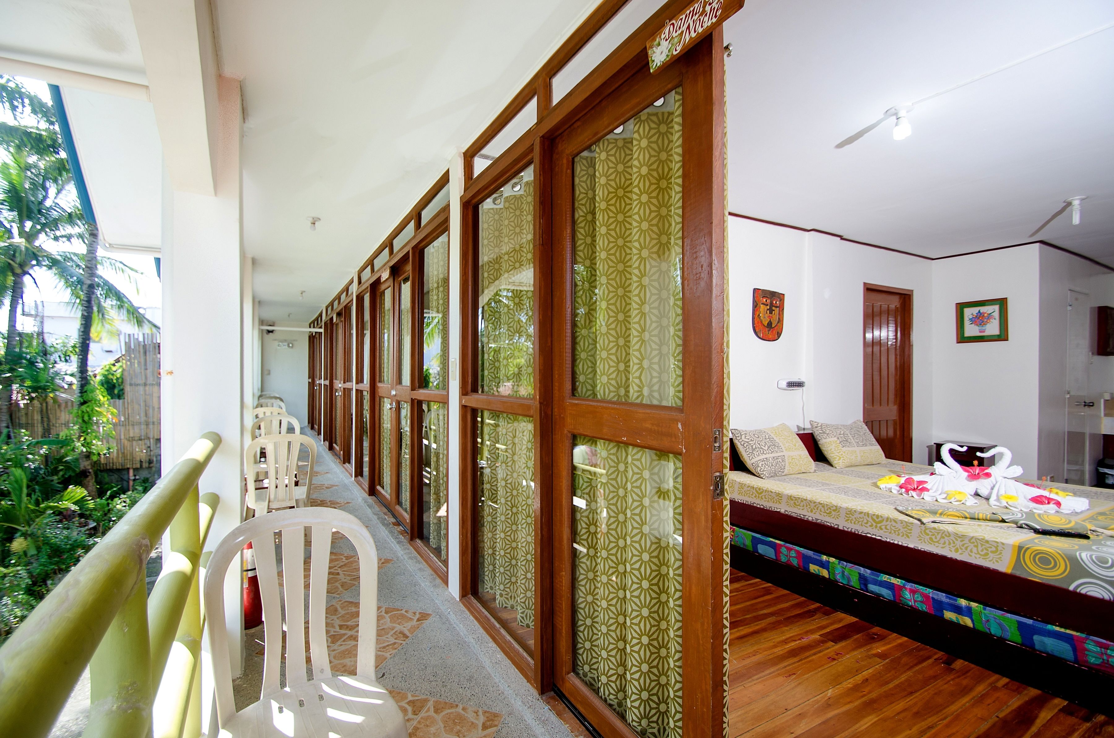 A room with a bed, a window with patterned curtains, a wooden door, and a view of the outdoors.