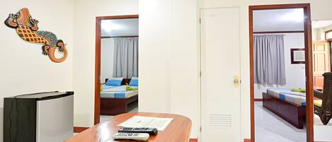 A hotel room with a wooden desk, a refrigerator, a bed with a blue and white bedspread, and a bathroom visible through an open door.