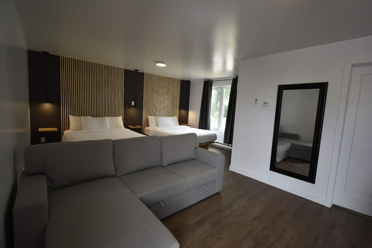 Moderni hotellihuone, jossa on harmaa kulmasohva, valkoisilla lakanoiden peitetty sänky, suuri peili ja ikkuna, jossa on verhot.