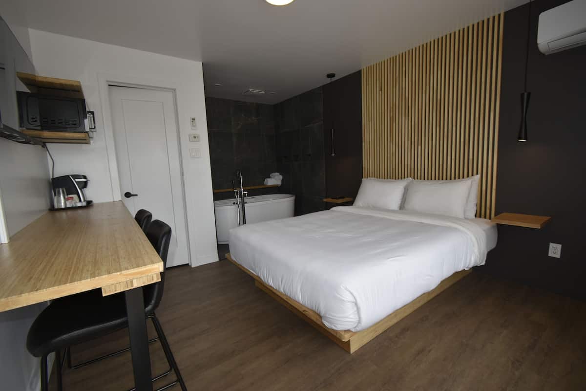 Moderni hotellihuone, jossa on puinen sänky, työpöytä tuolilla, pieni keittokomero ja suihkutila.