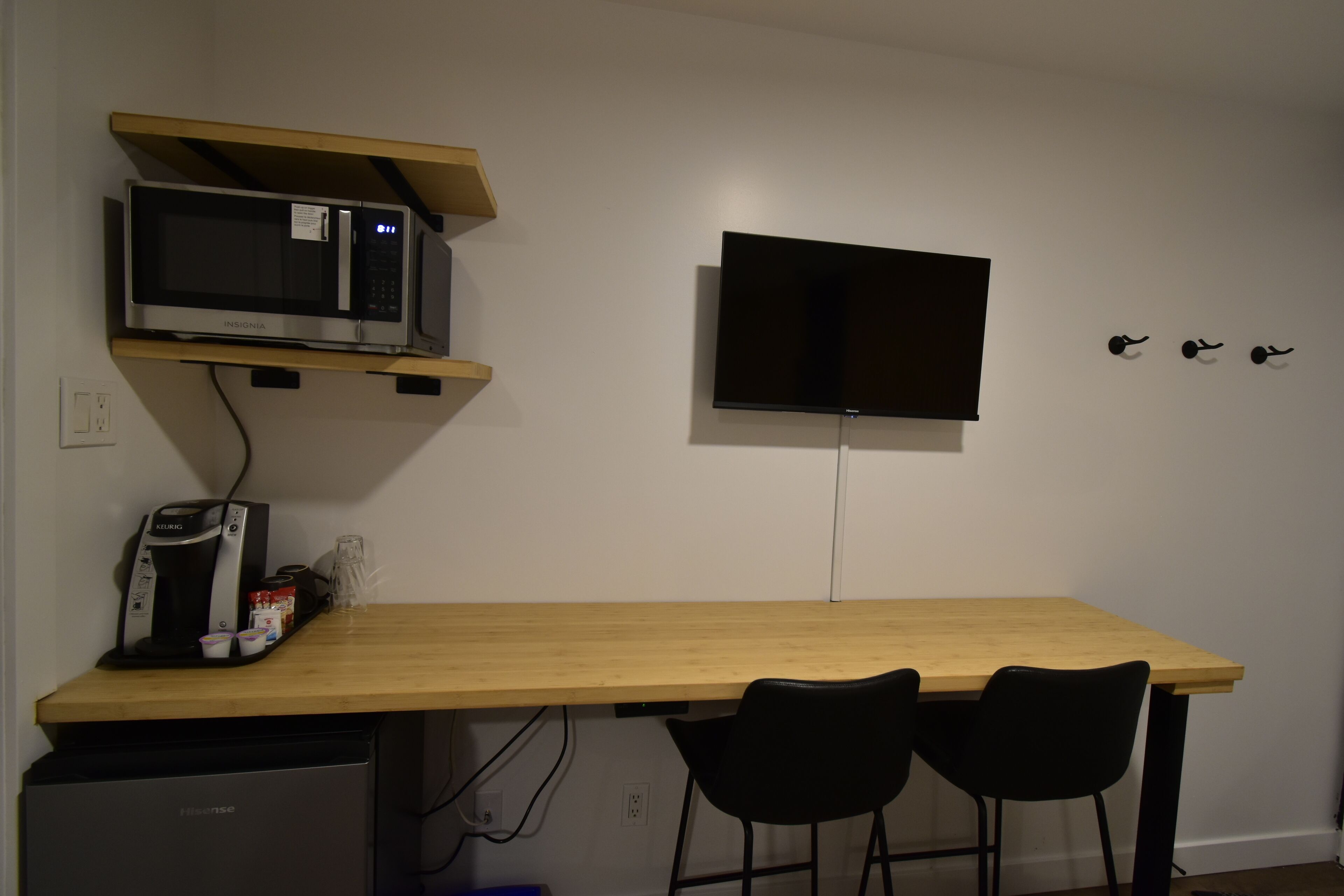 Un angolo cottura con forno a microonde, macchina del caffè e piccolo frigorifero. C'è una TV montata a parete e un tavolo di legno con due sedie.