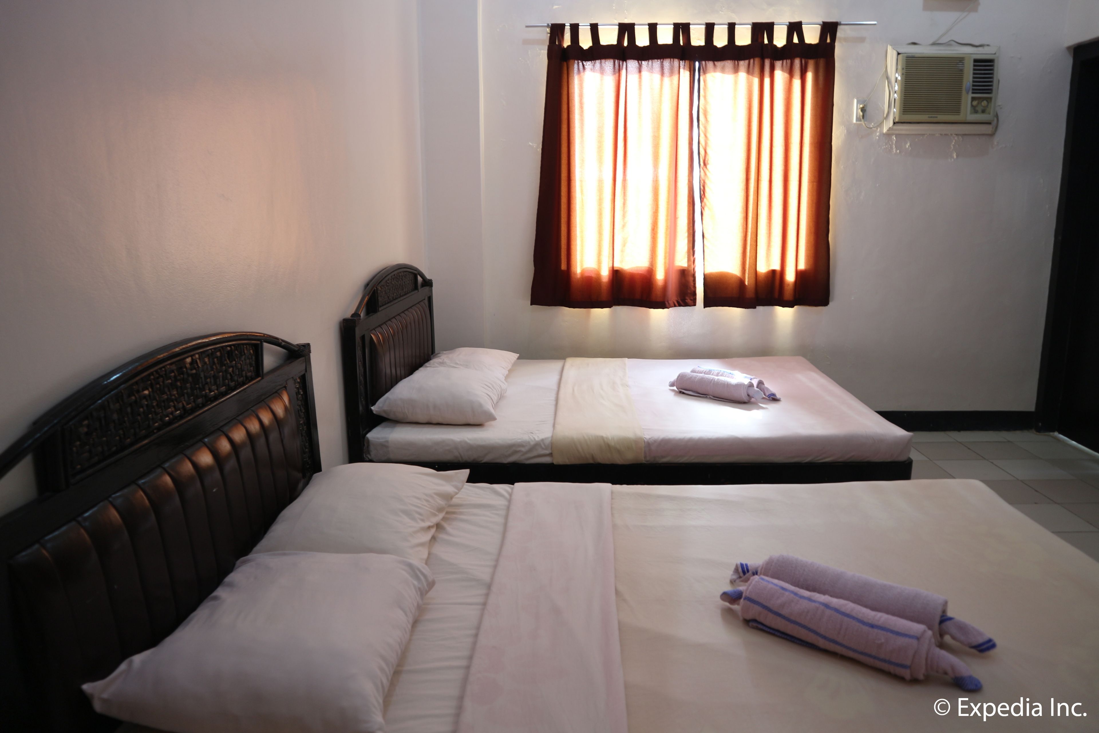A room with two single beds, a headboard, a window with curtains, an air conditioning unit, and a tiled floor.