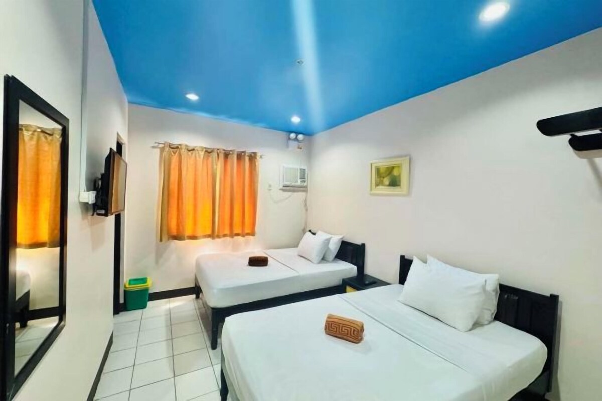A hotel room with two beds, a mirror, a window with curtains, a small air conditioning unit, and a picture on the wall.