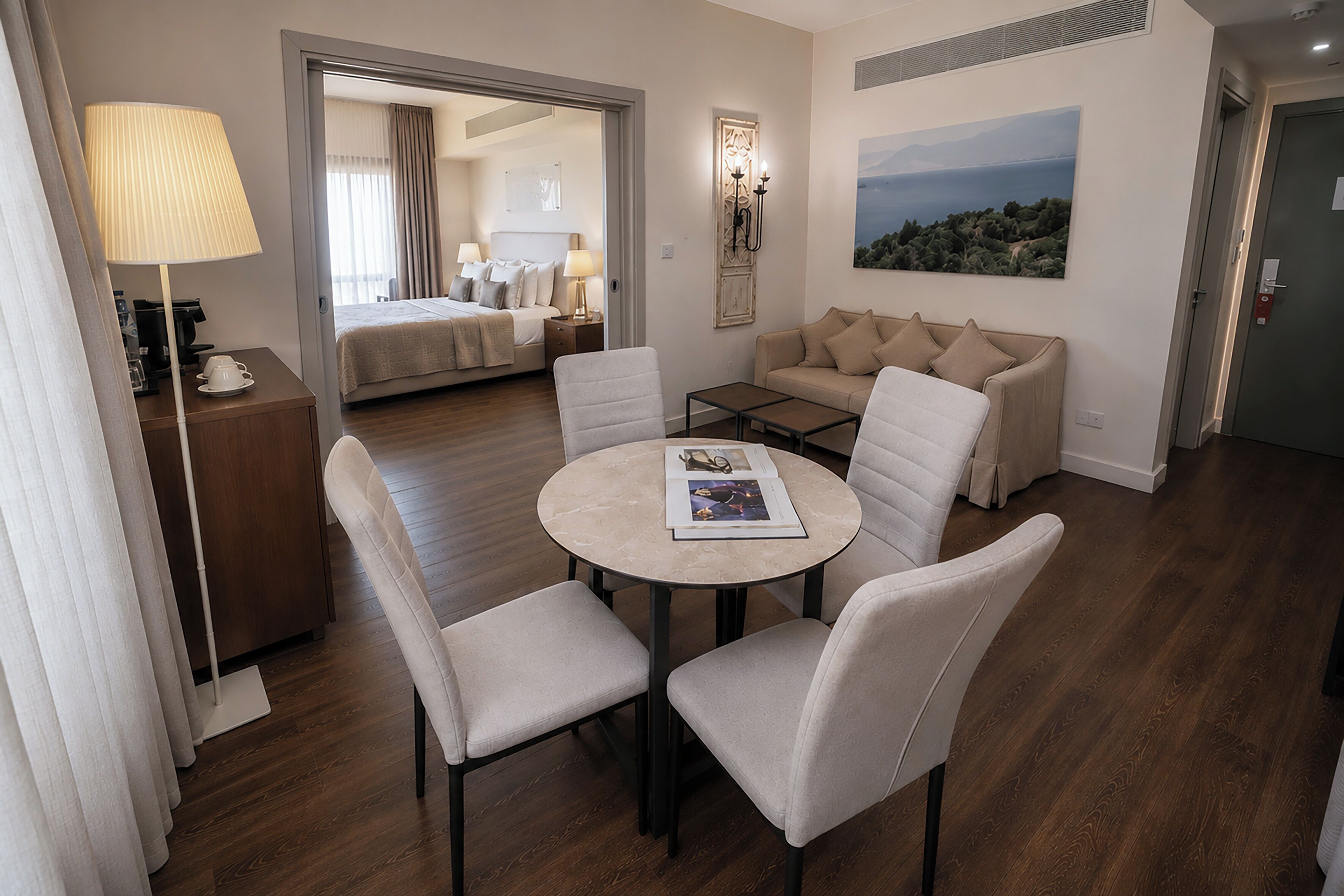 A hotel room with a bed, a sofa, a dining table with chairs, a lamp, and a painting on the wall.