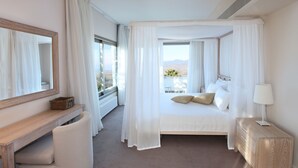 A hotel room with a bed, a desk, a chair, a bedside table, and a large window with curtains. - Droushia Heights (Droushia)