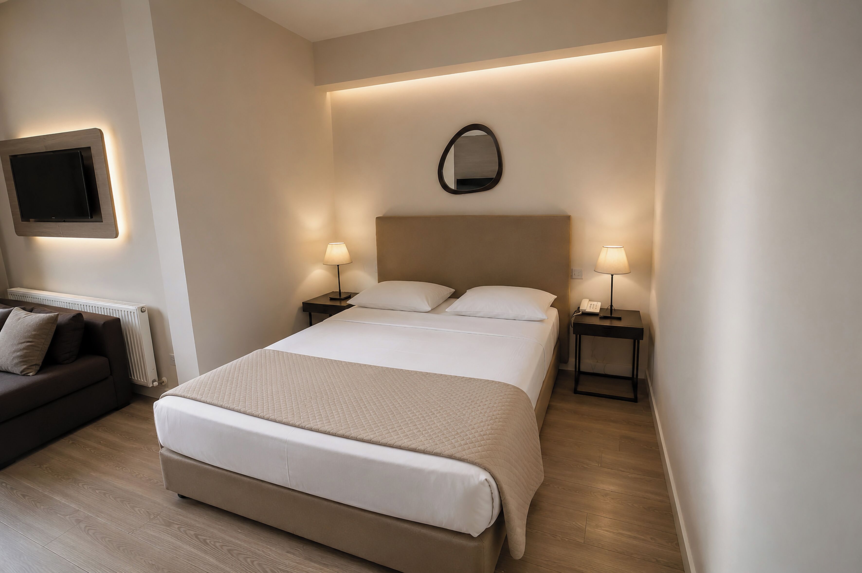 A hotel room with a large bed, two bedside tables with lamps, a flat-screen TV mounted on the wall, and a dark-colored sofa.