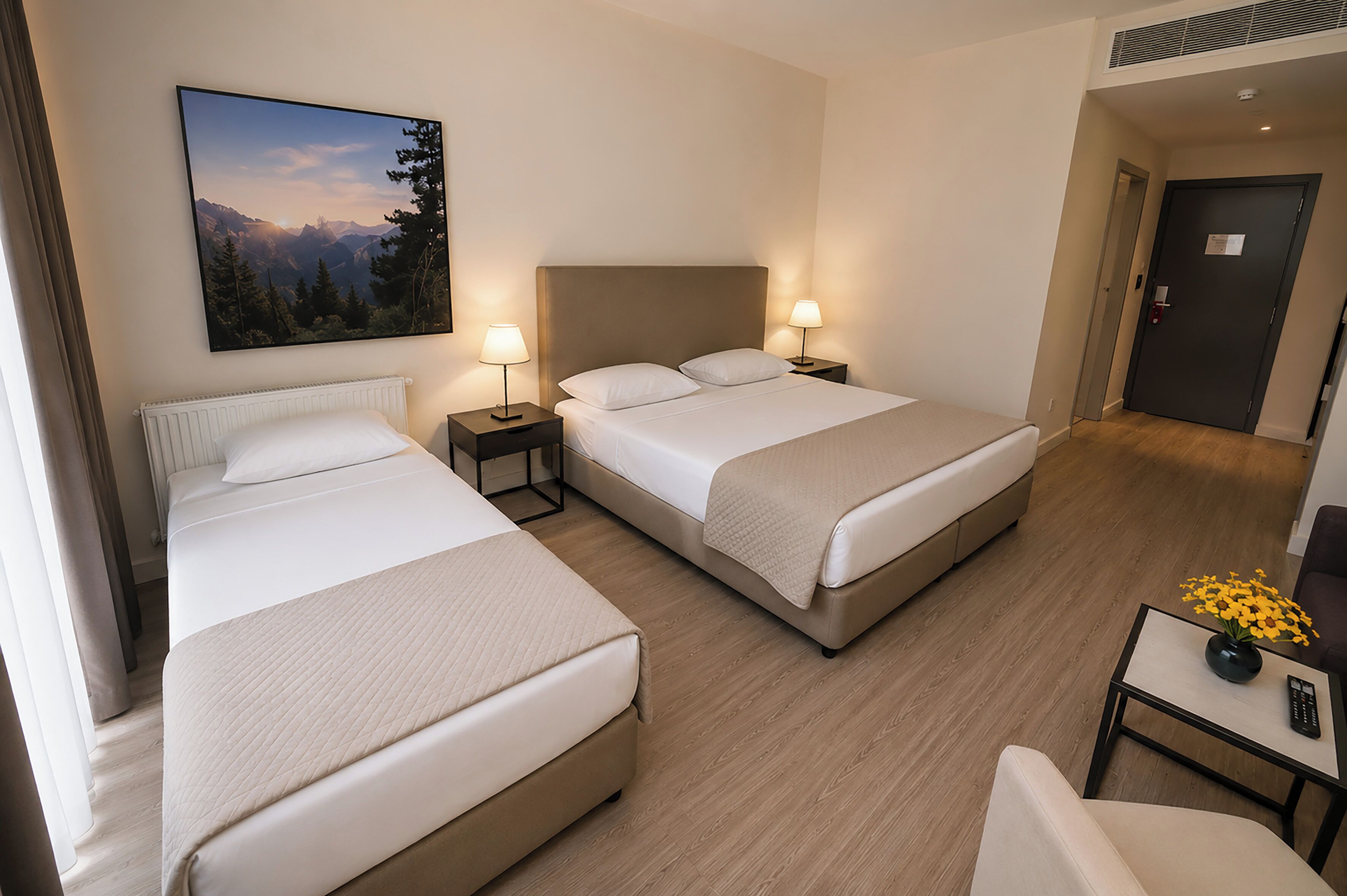 A hotel room with two beds, a painting of a mountain landscape, a small table with a vase of flowers, and a door.