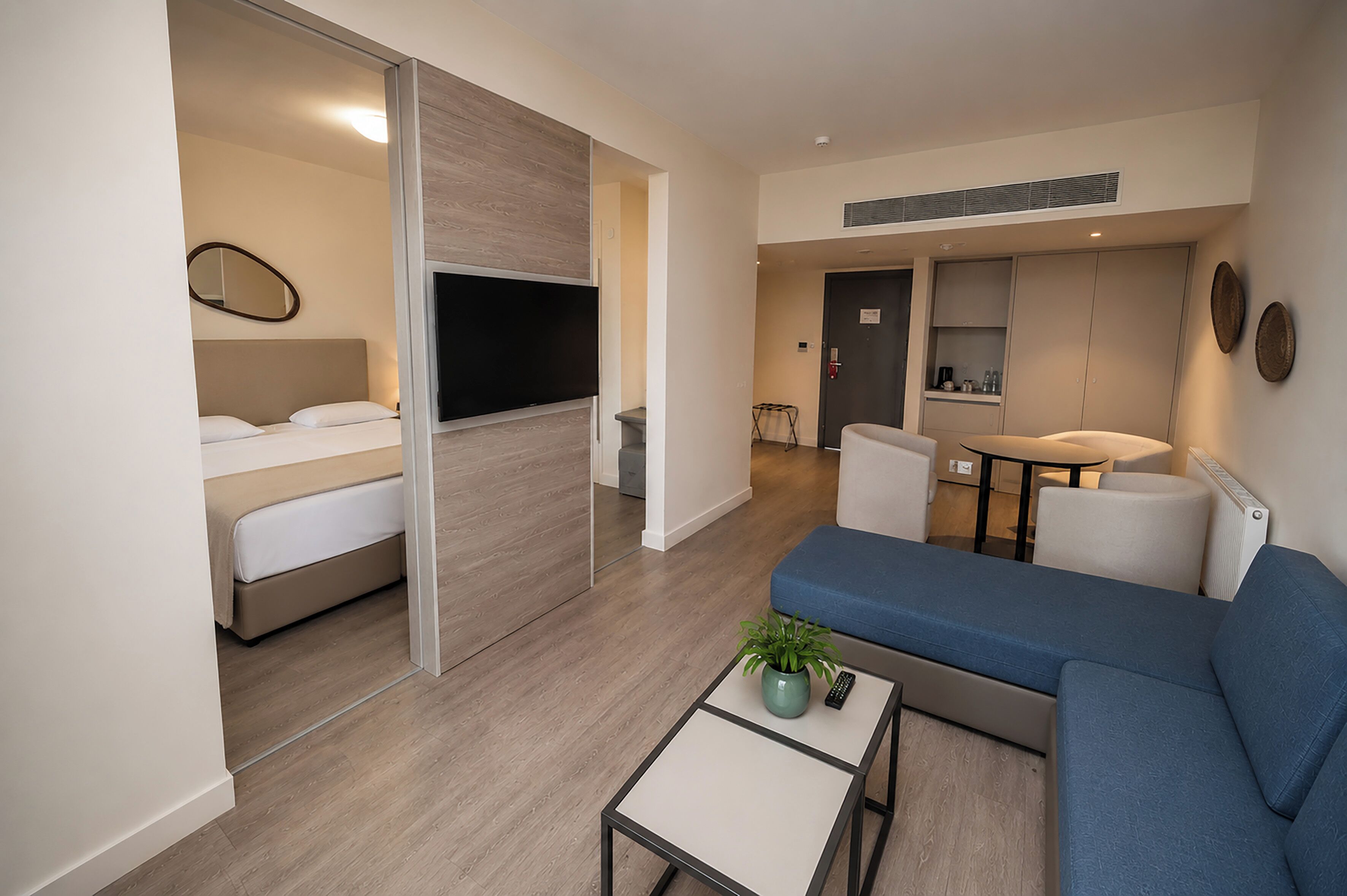 Una habitación de hotel moderna con una cama, un sofá, una mesita con una planta, un televisor y una kitchenette.