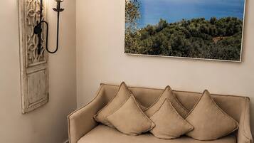 Un sofá color crema con cuatro cojines, una pequeña mesa cuadrada con un jarrón de flores amarillas, un aplique de pared y un cuadro grande con un paisaje.
