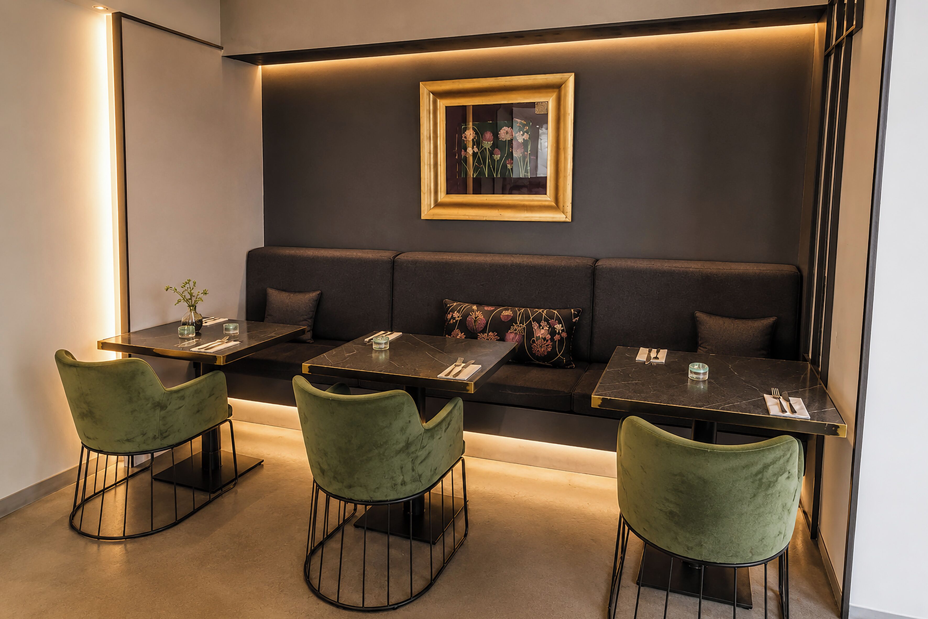 Un espace repas moderne avec un canapé de couleur sombre, des chaises recouvertes d’un tissu vert et une œuvre d’art florale encadrée accrochée au mur.