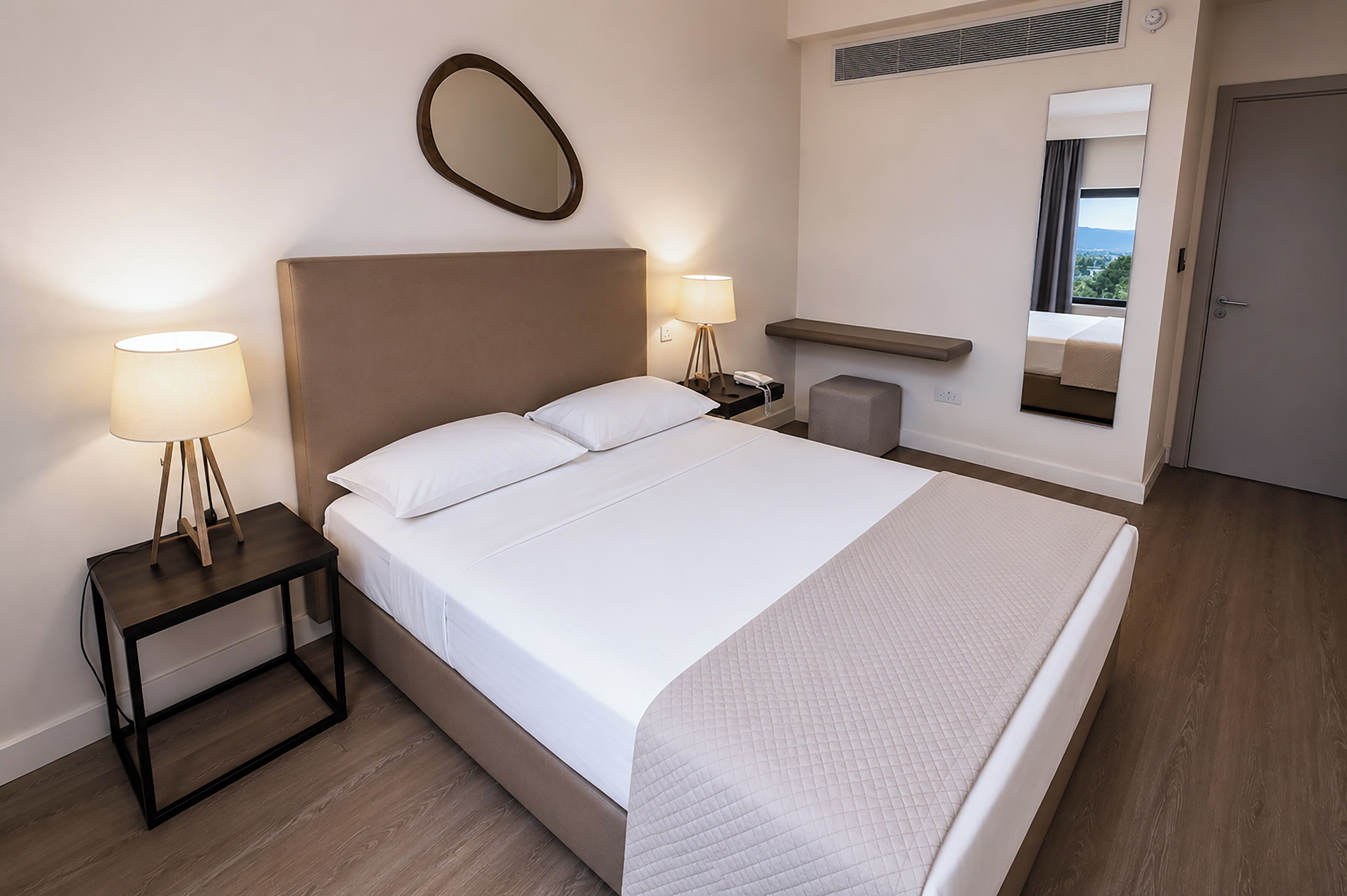 A neatly made bed with white linens, a bedside table with a lamp, a small stool, a mirror, and a view of the outdoors through a window.