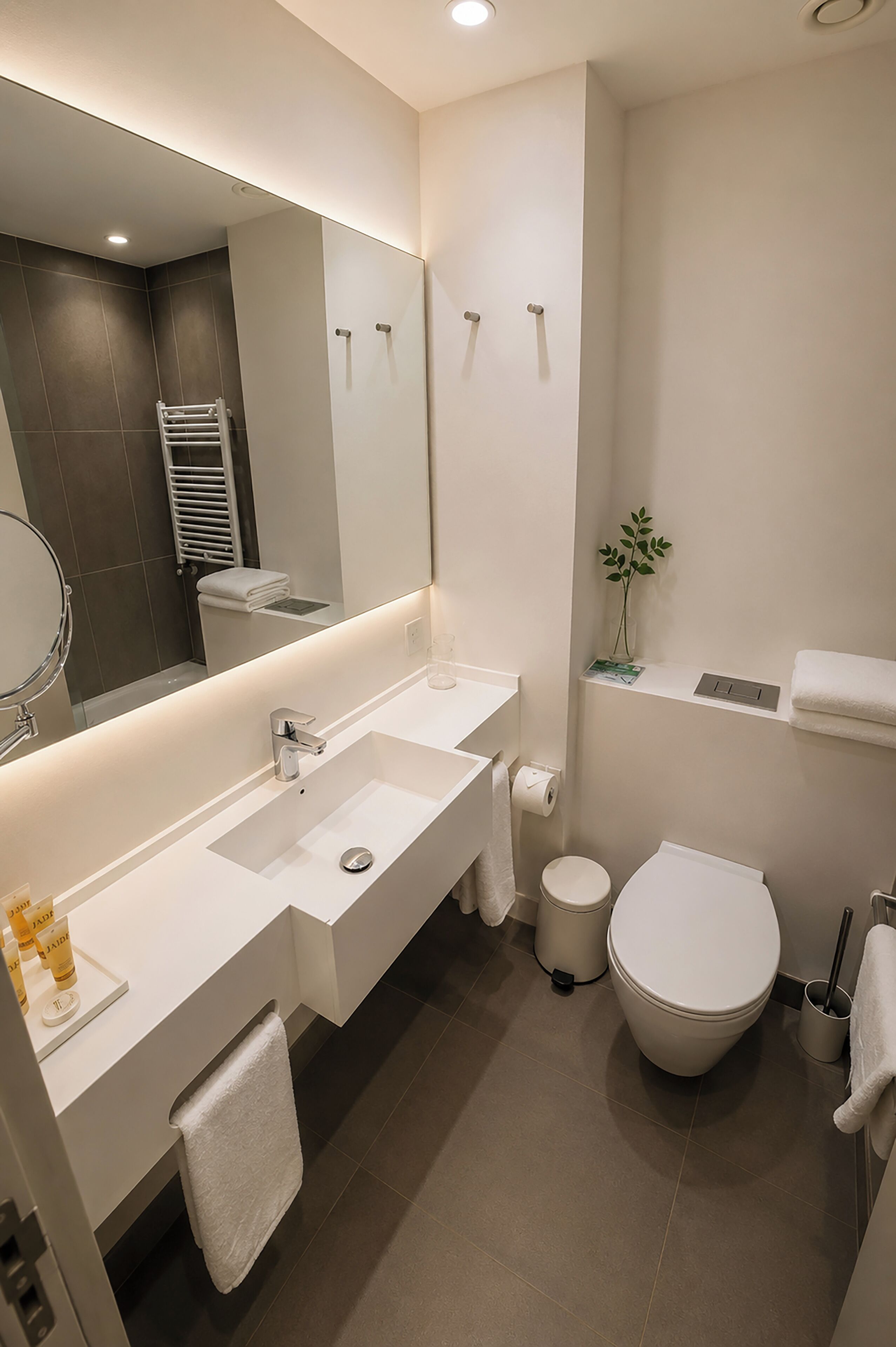 Une salle de bain moderne avec un grand miroir, deux lavabos, des toilettes et une petite plante posée sur une étagère.