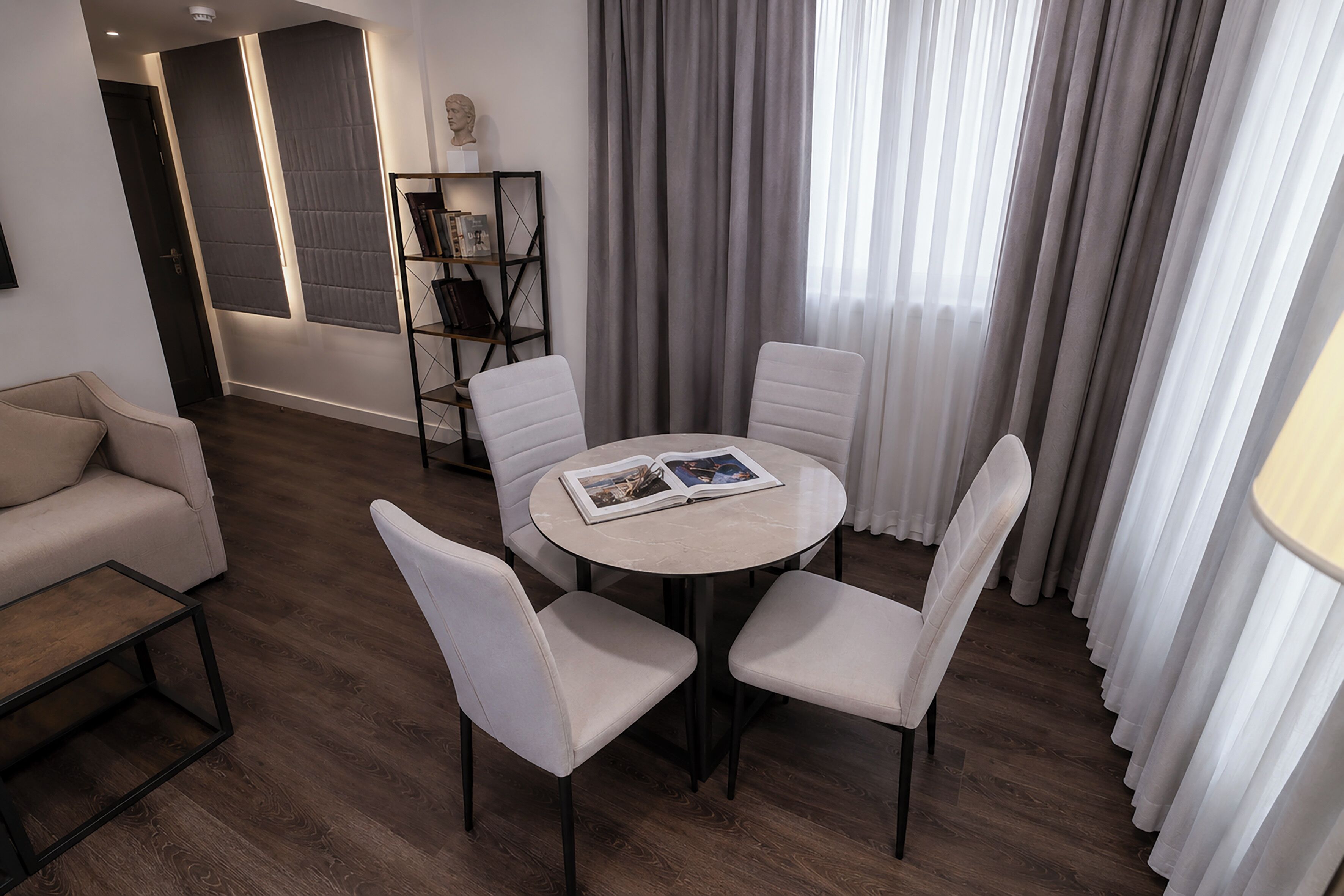 A modern living room with a round table, four white chairs, a sofa, a bookshelf, and a large window with curtains.