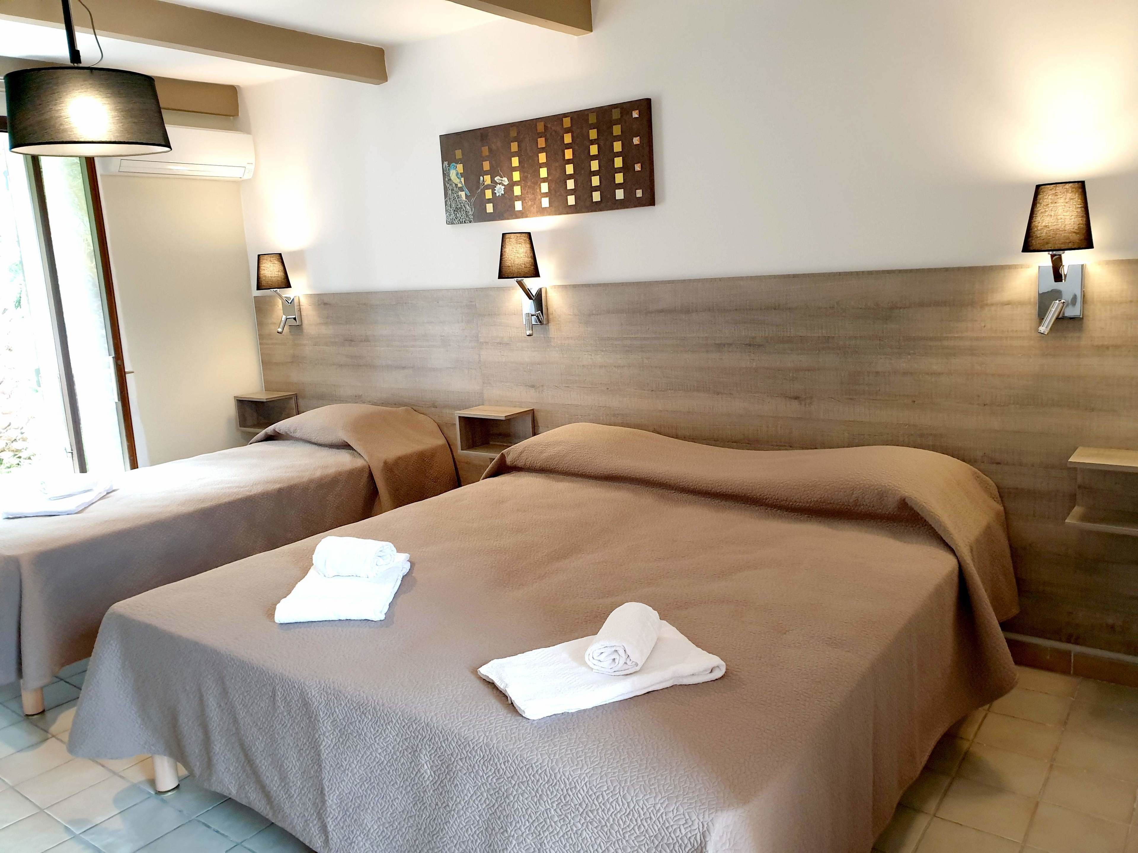 Habitación de hotel con dos camas, cada una con una colcha color beige y una toalla enrollada. Hay lámparas fijadas a la pared y una pieza decorativa sobre las camas.