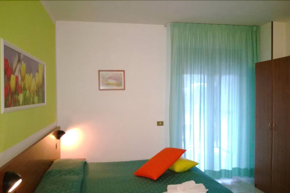 Una habitación con una cama, una pared verde, un cuadro, un marco en la pared y una ventana con cortinas.