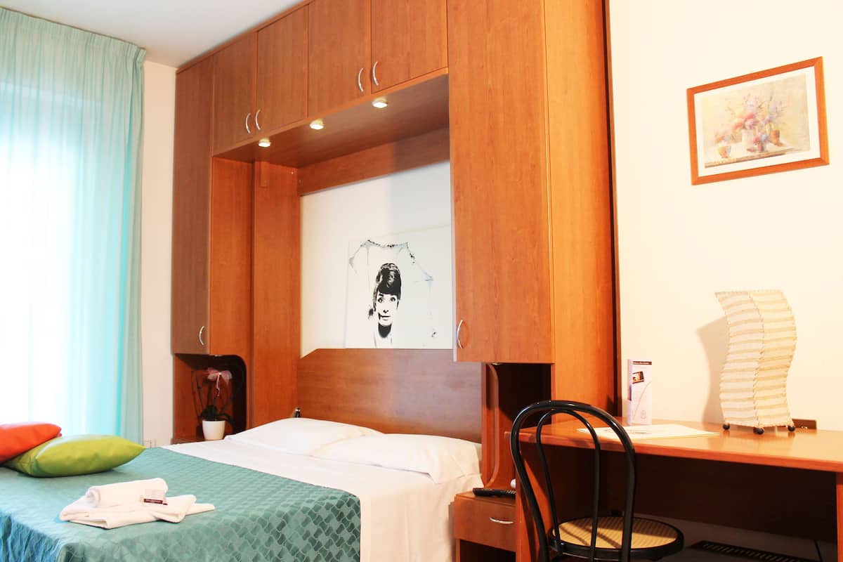 Habitación de hotel con cama, escritorio, silla, planta en maceta y un cuadro en la pared.