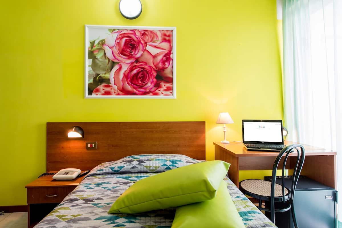 Una habitación de hotel con una cama, un escritorio con una computadora portátil, un teléfono y un cuadro de rosas en la pared.