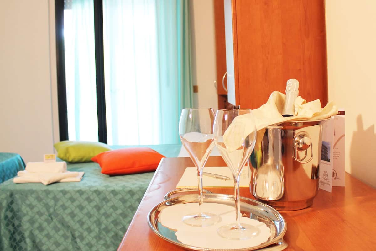 Habitación de hotel con una cama, un atril con vasos y una botella, y una ventana con cortinas.