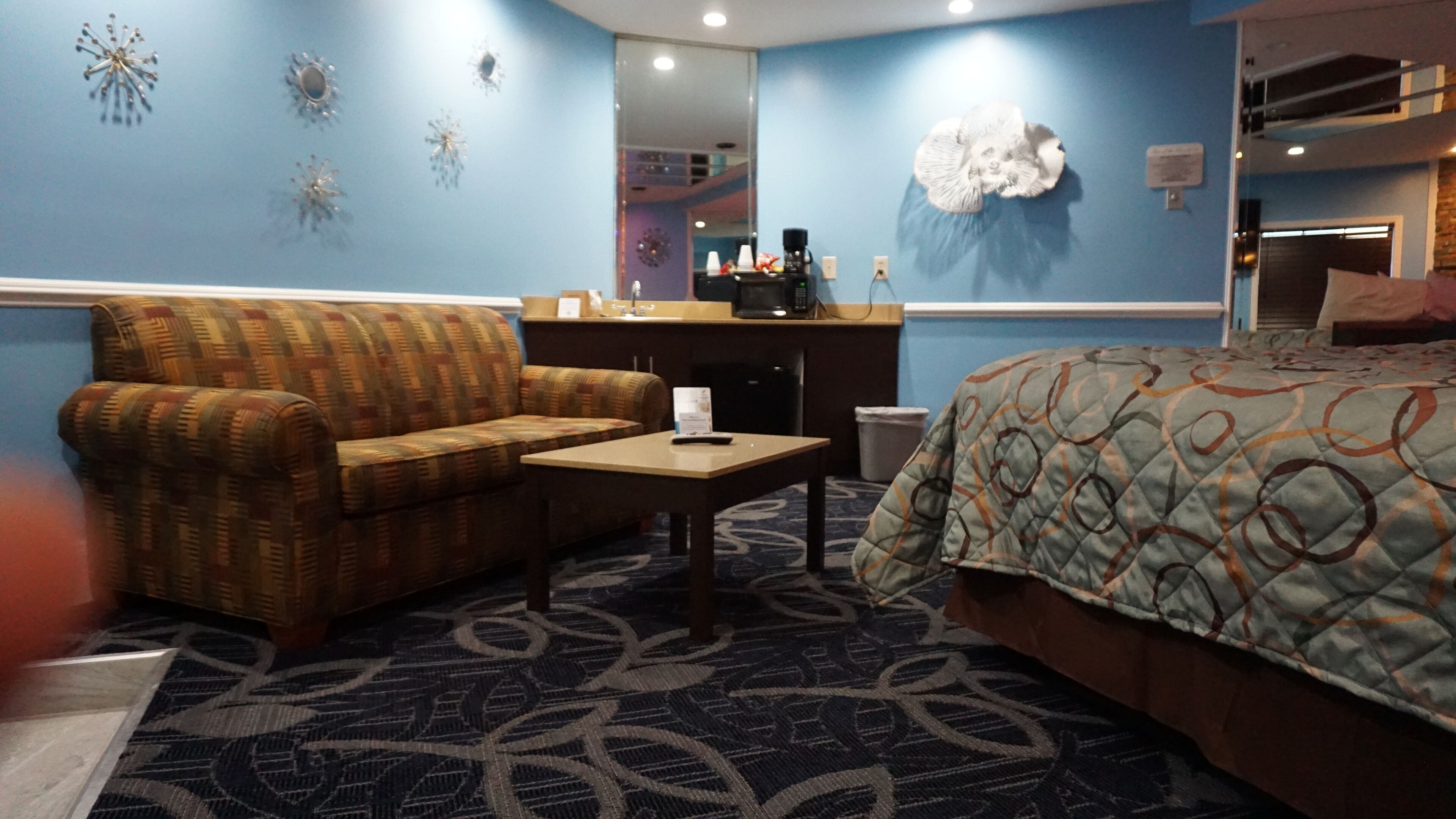 A hotel room with a sofa, a small table, a bed with a patterned blanket, a coffee machine, and decorative wall art.