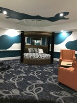 Un dormitorio con una cama con dosel, un banquillo redondo, una silla y una alfombra estampada.