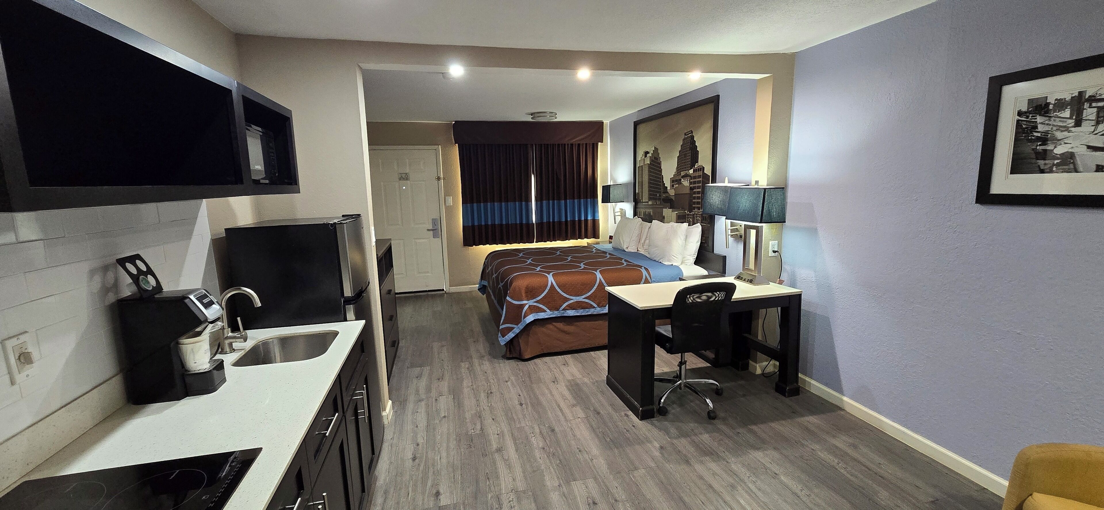 A hotel room with a kitchenette, a bed with a patterned bedspread, a desk with a chair, and a framed picture on the wall.