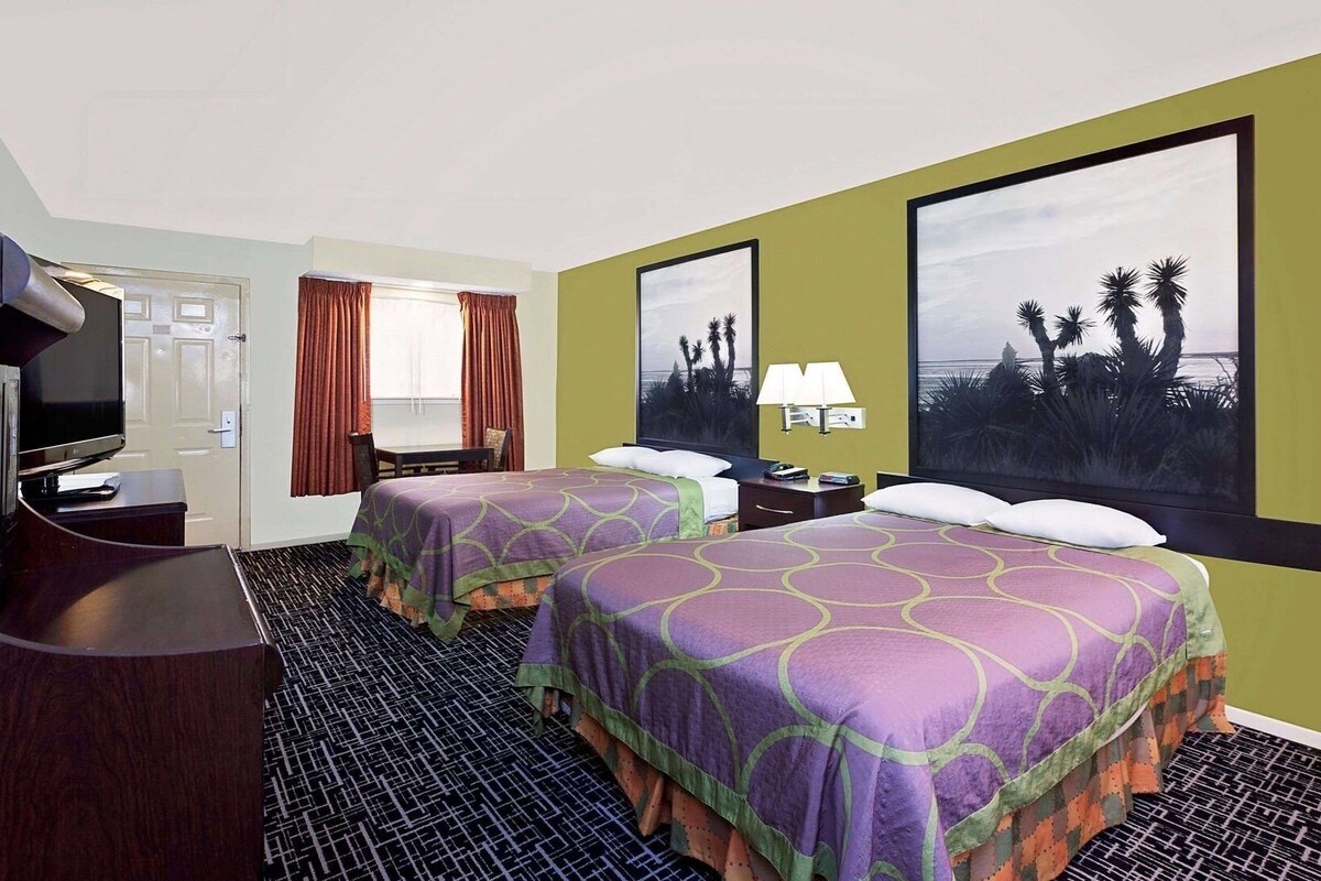 A hotel room with two beds, a TV, a desk, a window with curtains, and a large painting of a beach scene.