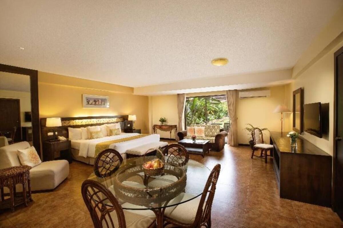 Een hotelkamer met een bed, een eettafel met stoelen, een bank, een televisie en een raam met uitzicht op groen.
