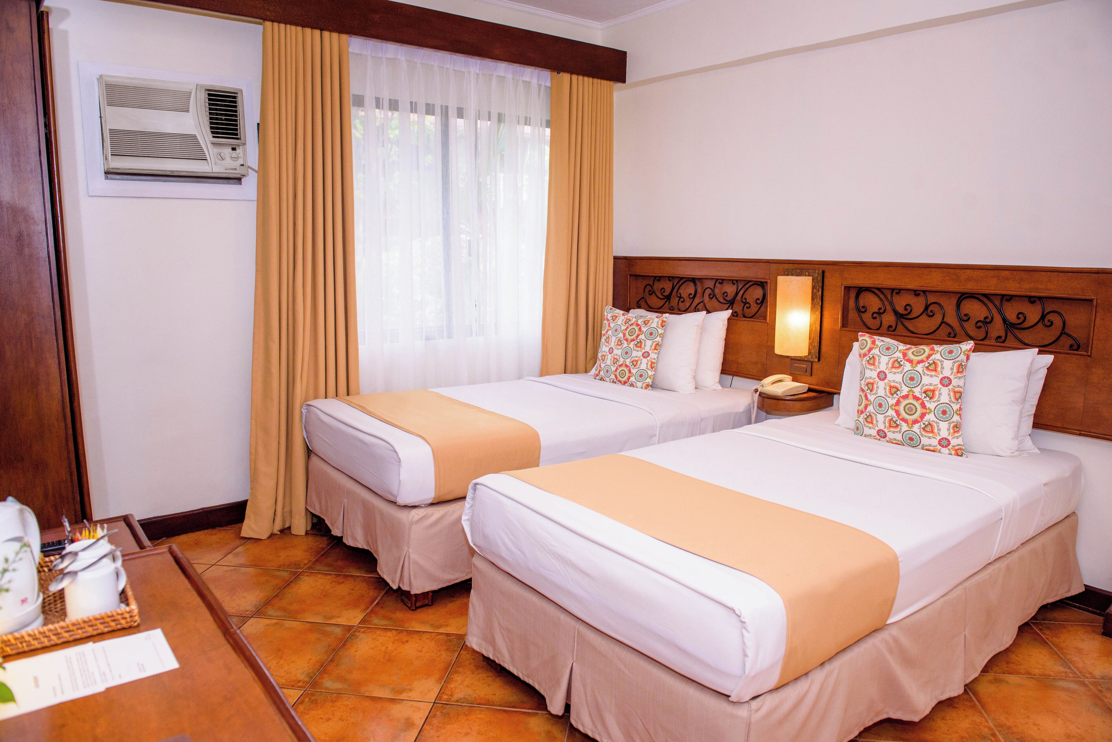 A hotel room with two beds, a bedside table with a lamp, an air conditioning unit, and a window with curtains.