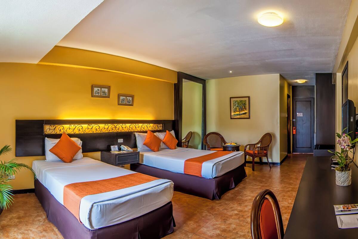 Een hotelkamer met twee bedden, een bureau met spiegel, een potplant en een deur naar een andere kamer.