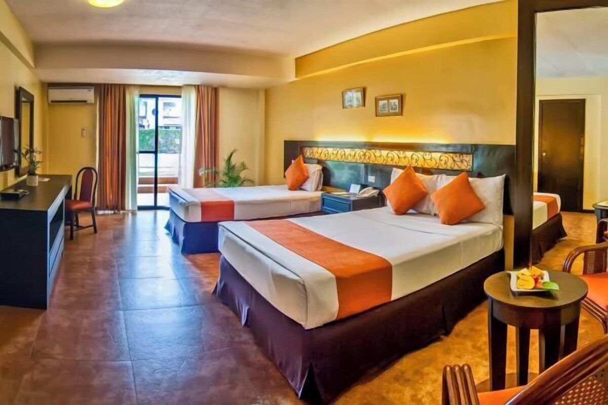 Quarto de hotel com duas camas, uma escrivaninha, uma cadeira e uma mesinha.