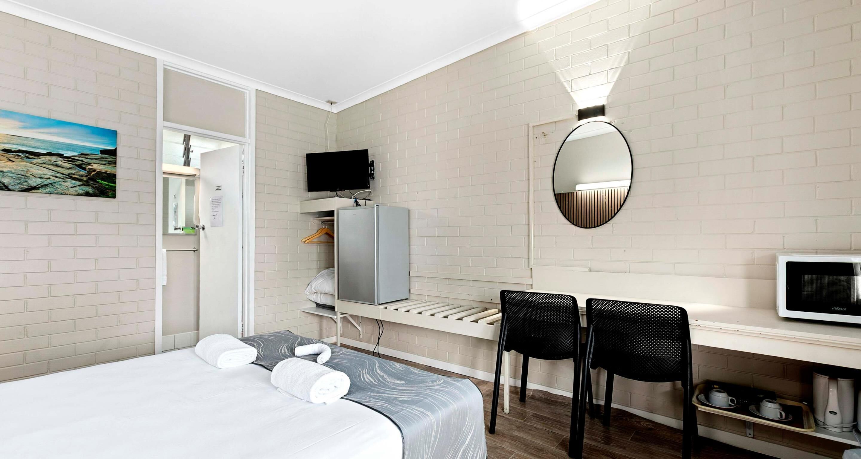 A hotel room with a bed, a desk with a computer and a TV, a mirror, and a small kitchenette.