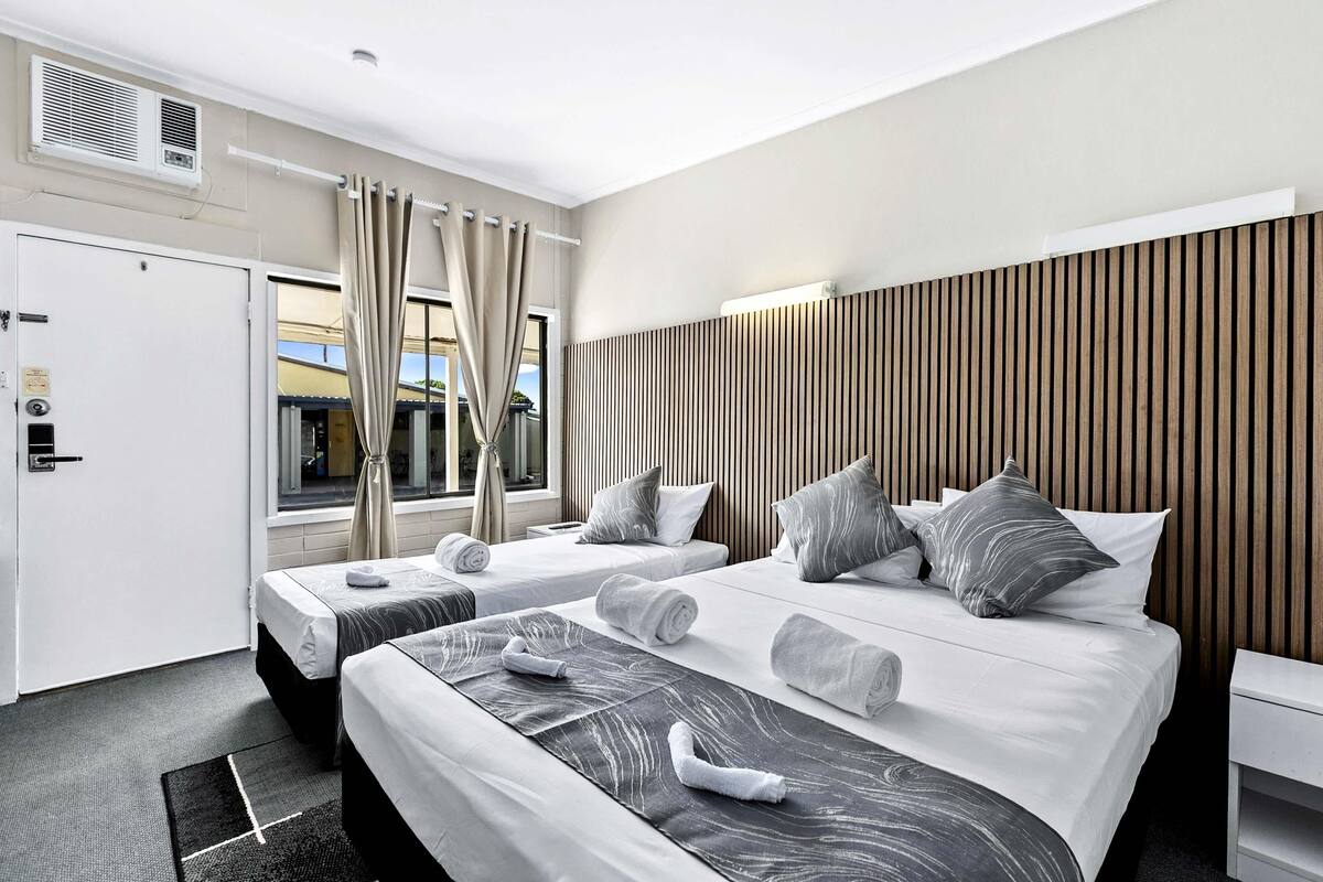 Hotellihuone, jossa on kaksi sänkyä, ikkuna, jossa on verhot, puupanelilla verhoiltu seinä ja valkoinen ovi.