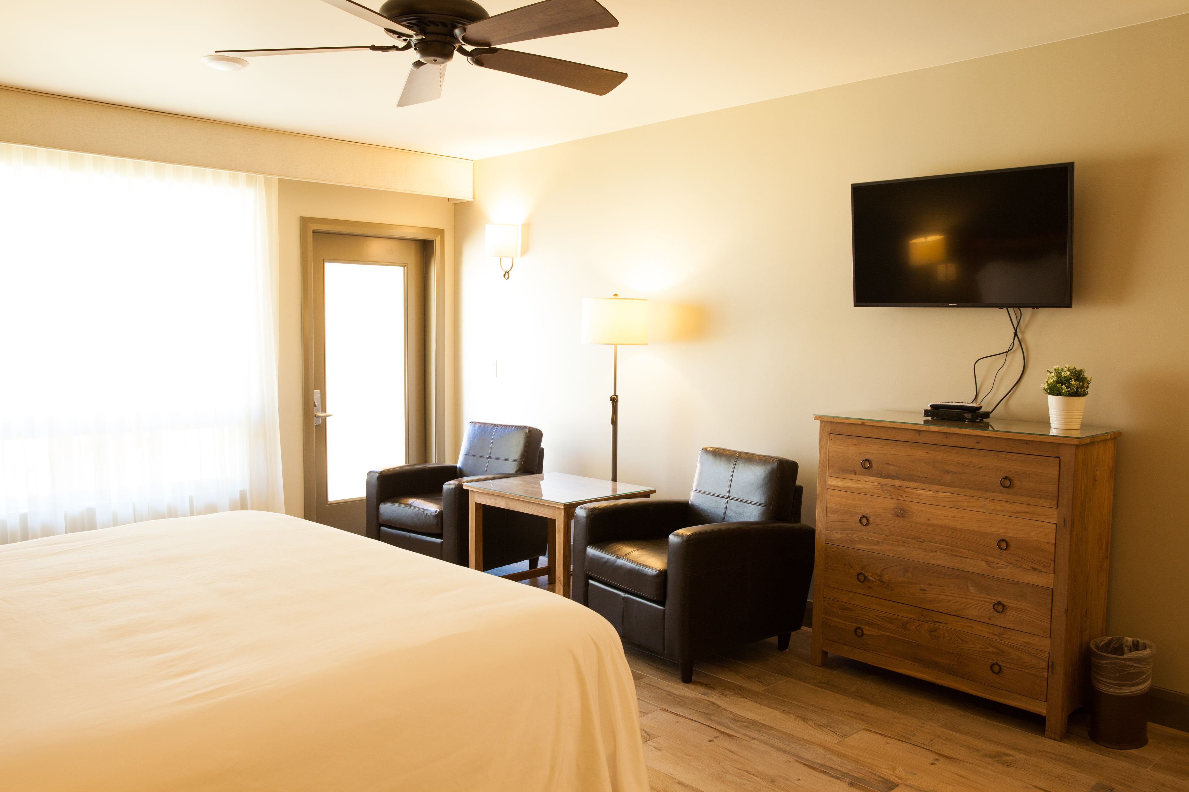 A hotel room with a bed, two chairs, a table, a dresser, a television, and a ceiling fan.