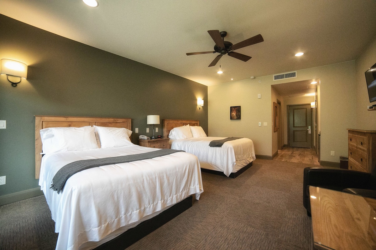 A hotel room with two beds, a ceiling fan, wall sconces, a flat-screen TV, and a wooden dresser.