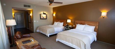 A hotel room with two beds, a desk, a chair, a ceiling fan, and wall-mounted lamps.