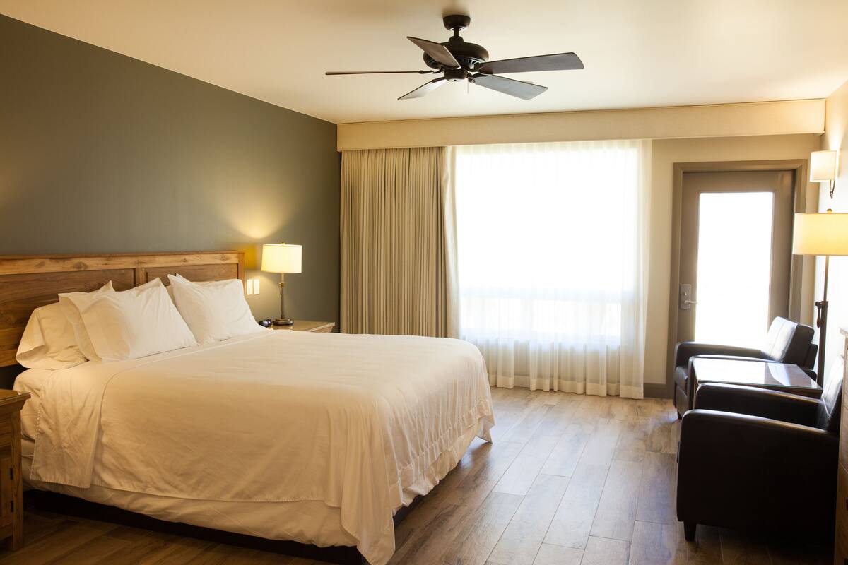A hotel room with a large bed, a ceiling fan, a desk with a chair, and a window with curtains.