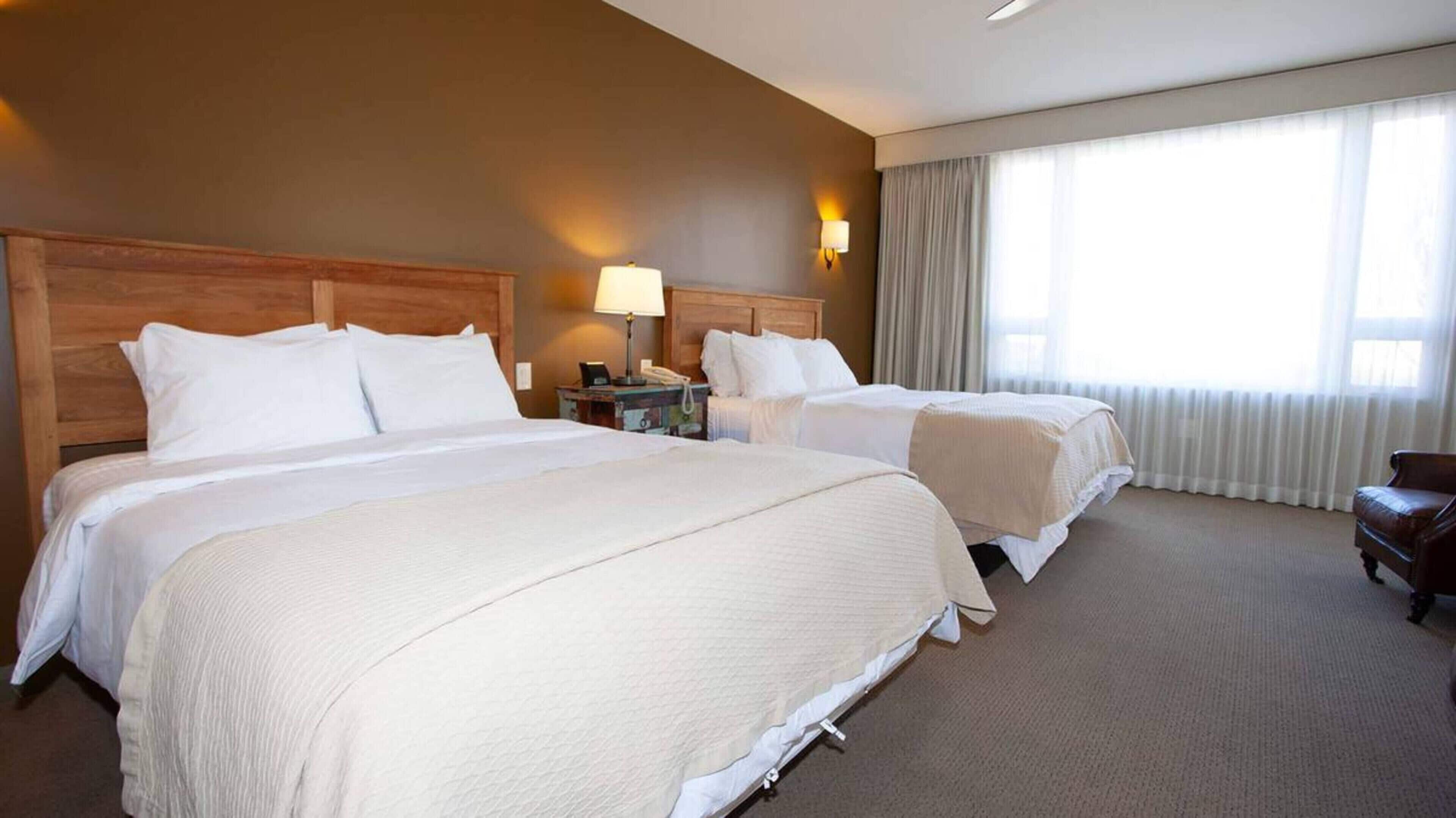 Habitación de hotel con dos camas, cabecero de madera, mesita de noche con lámpara, teléfono y ventana con cortinas.