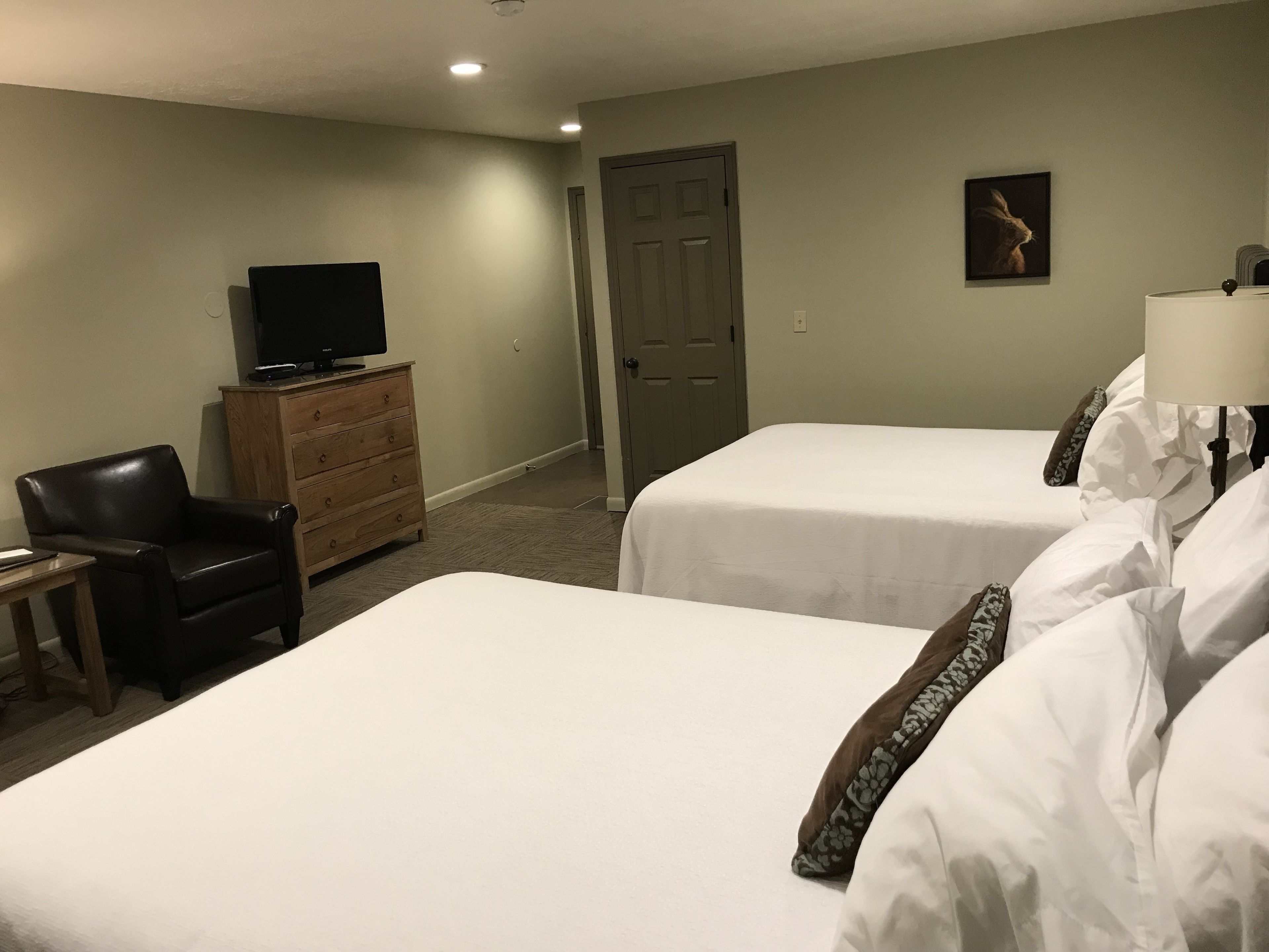 Ein Hotelzimmer mit zwei Betten, einem Fernseher auf einem Schminktisch, einem Sessel und einer Tür.