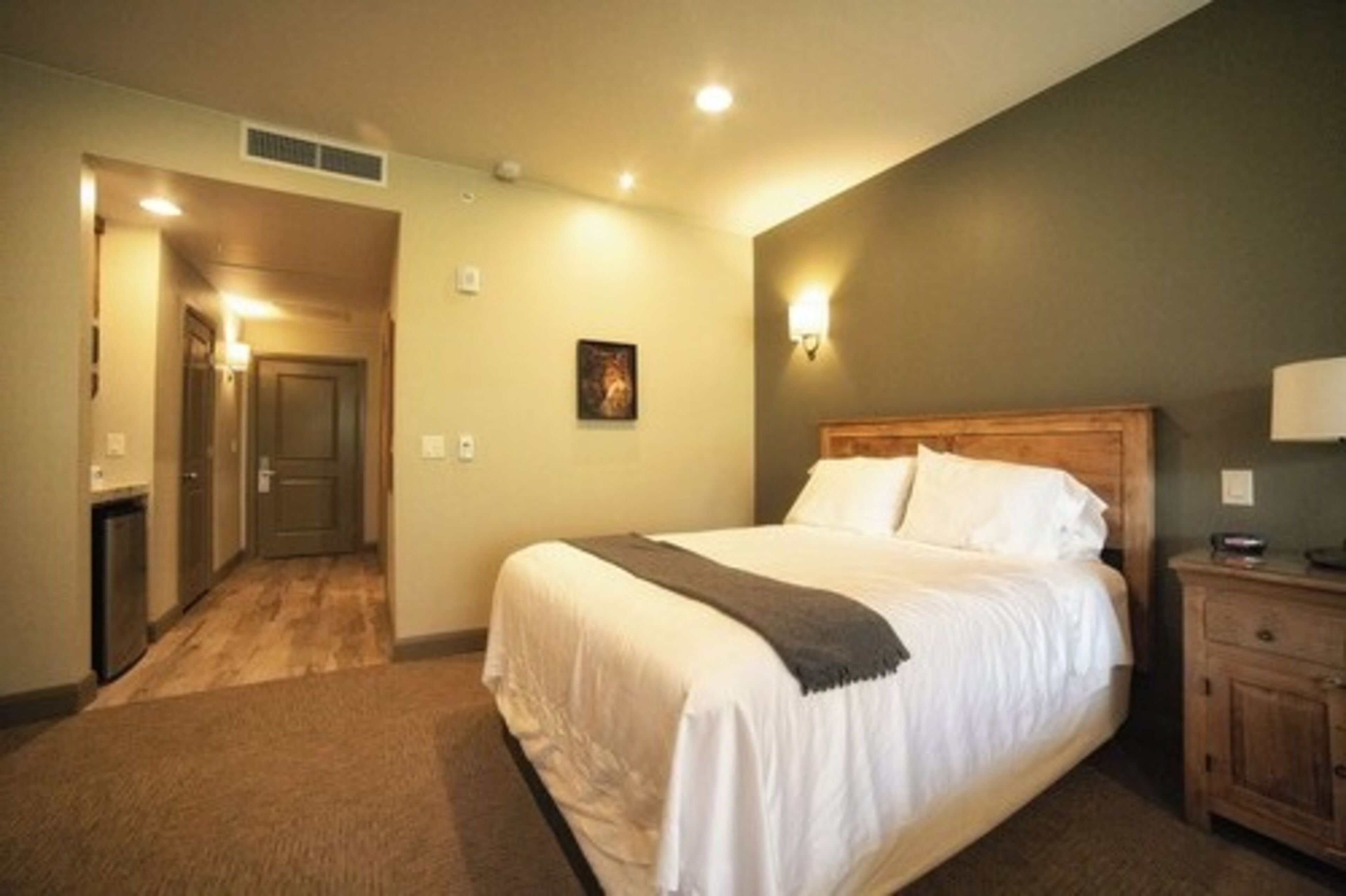 Une chambre d’hôtel avec un grand lit, une table de chevet en bois, une lampe fixée au mur et une porte donnant sur un couloir.