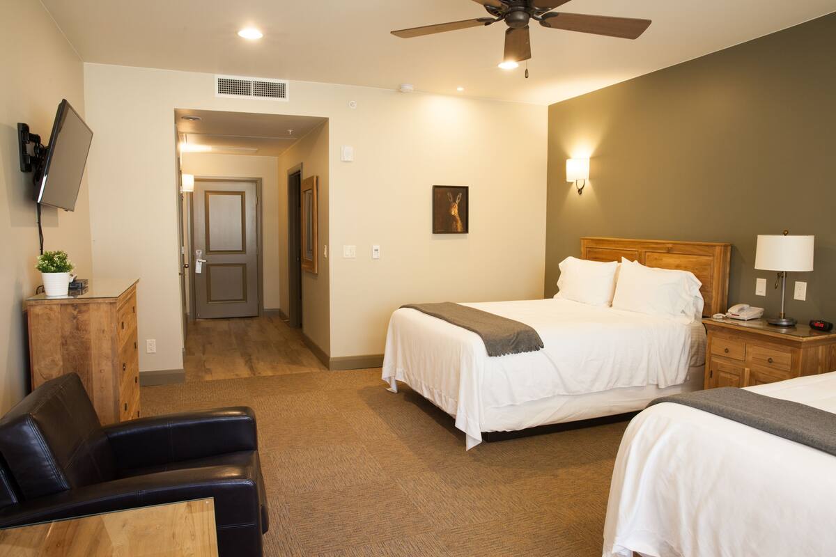 A hotel room with two beds, a desk, a chair, a TV, a ceiling fan, and a door leading to another room.