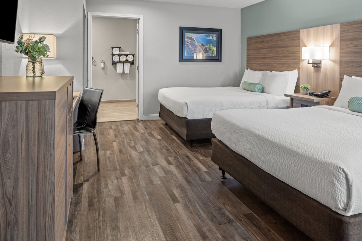 Ein Hotelzimmer mit zwei Betten, einem Schreibtisch und einem Stuhl. Es gibt einen Holzboden, eine an der Wand befestigte Uhr und ein gerahmtes Bild.