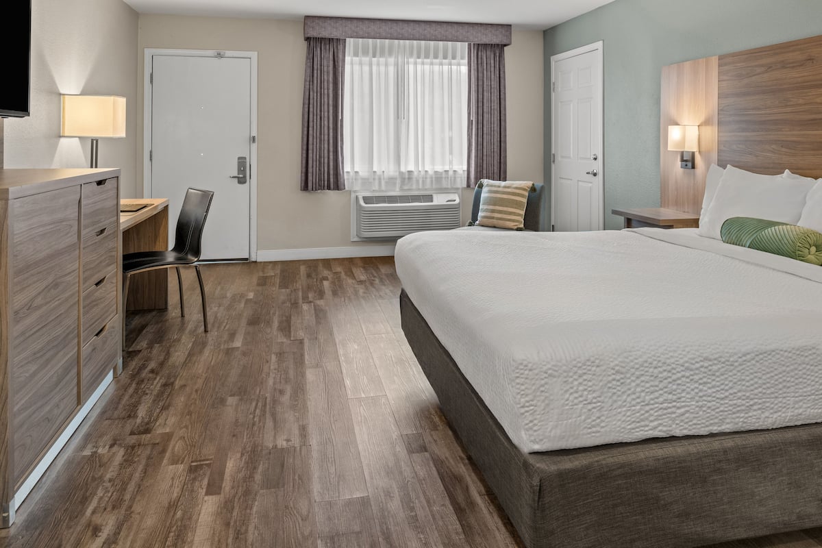 Ein Hotelzimmer mit Bett, Schreibtisch, Stuhl, Kleiderschrank, einem Fenster mit Vorhängen und einer Wandlampe.