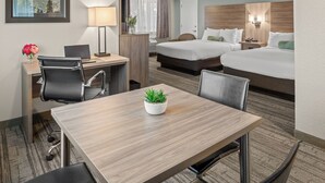 A hotel room with a desk, two beds, a chair, a table with a plant, a lamp, and a view of mountains. - Yosemite Westgate Lodge (Groveland)
