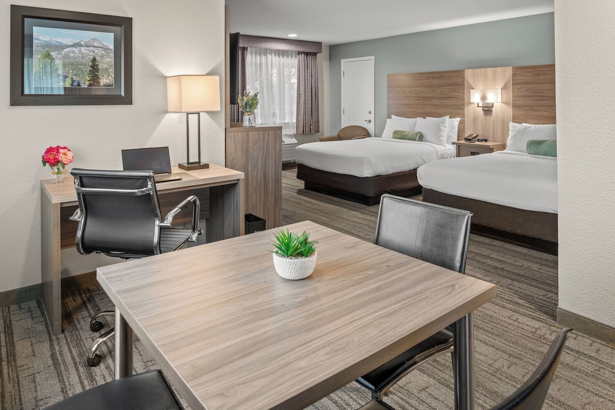 Ein Hotelzimmer mit Schreibtisch, zwei Betten, einem Sessel, einem Tisch mit Pflanze, einer Lampe und Blick auf Berge.